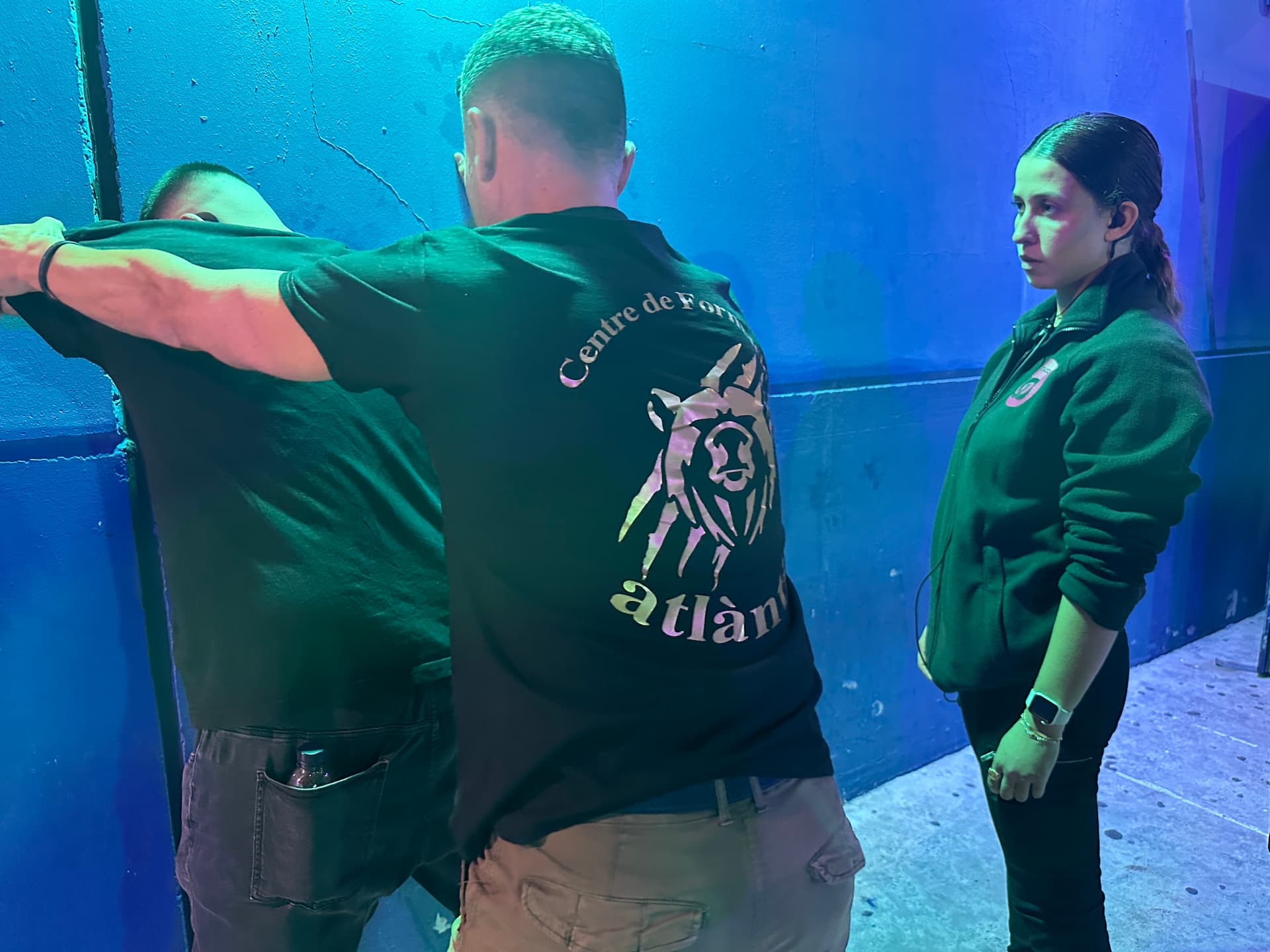 Dos personas realizando un control de seguridad a un hombre contra una pared azul, con una mujer observando de cerca.